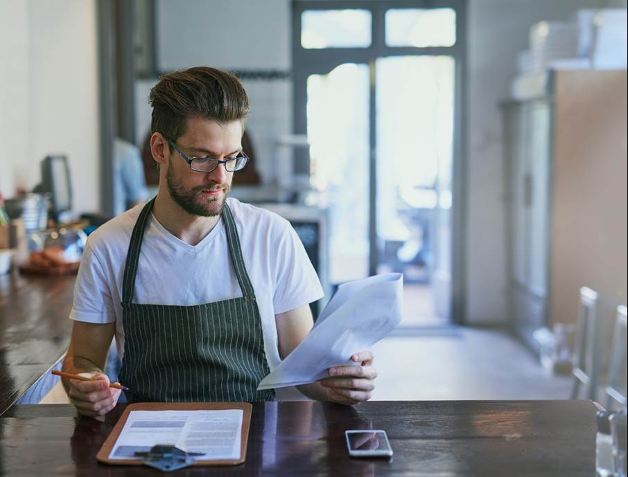 En man med glasögon och iklädd förkläde och beskrivs som barista, sitter vid ett bord och skriver anteckningar i ett block. 